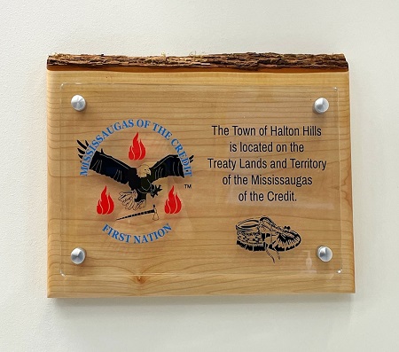 Wooden wall plaque with a clear acrylic front panel. On the left, a circular emblem shows an eagle with outstretched wings holding a feather, surrounded by three red flames and the text ‘Mississaugas of the Credit First Nation.’ On the right, engraved text reads: ‘The Town of Halton Hills is located on the Treaty Lands and Territory of the Mississaugas of the Credit.’ Below the text is an illustration of traditional moccasins. The plaque has four silver mounting bolts and a bark‑edged top